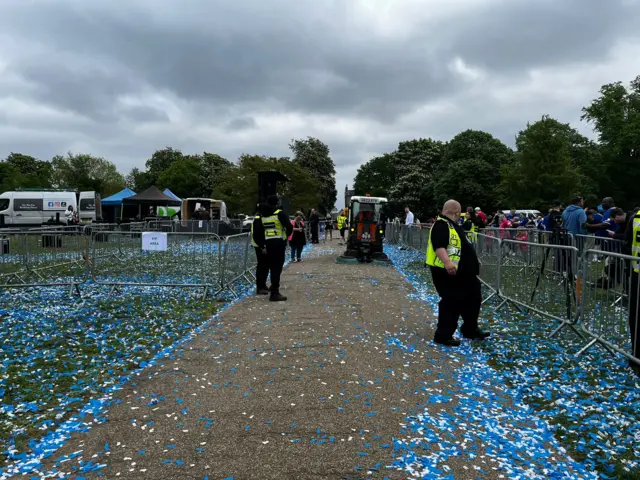 Confetti and streamers on the floor at Christchurch Park. There are security guards around metal fences.