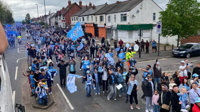 Fans with flags and banners celebrate alongside the bus