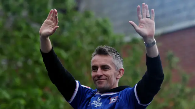 Kieran McKenna with his arms up on the Ipswich Town parade bus. He is wearing a blue football shirt and has his arms in the air.