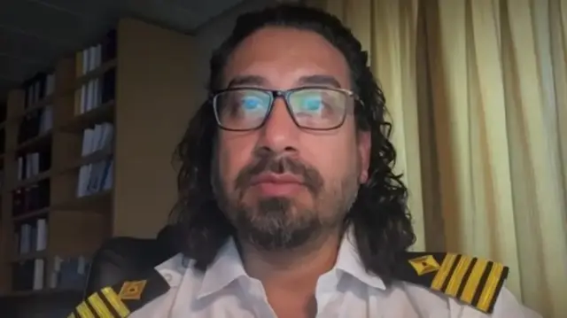 Captain Ramoon Kapoor sits inside a ship, with a drawn curtain and shelving unit visible behind him
