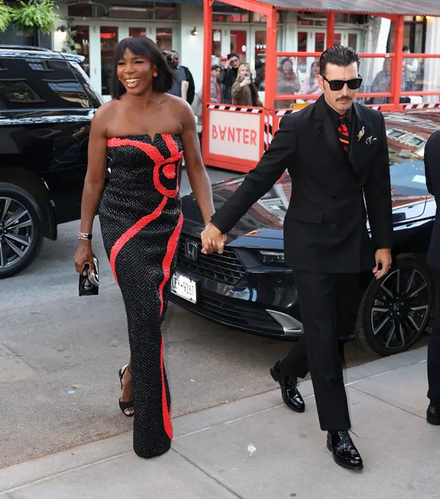Venus Williams, wearing a floor-length black glittery dress with a red swoop across it, holding hands with Pretti, wearing a black suit and red patterned tie