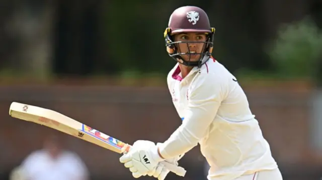 Josh Thomas holds his bat and wears a helmet as he plays cricket for Somerset