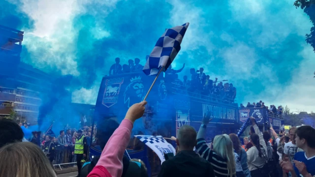 Fans line the way as Ipswich Town buses pass through. There are flags and blue flares have been let off