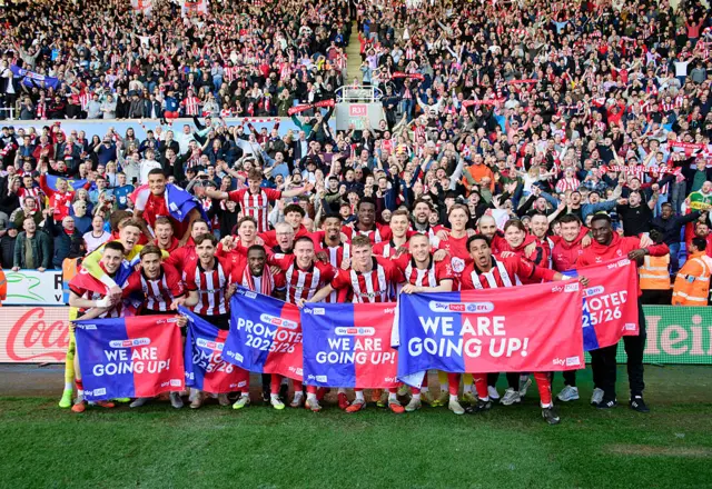 Lincoln City players wearing red and white striped shirts stand on the pitch in front of their jubilant fans holding red and blue promotion banners.