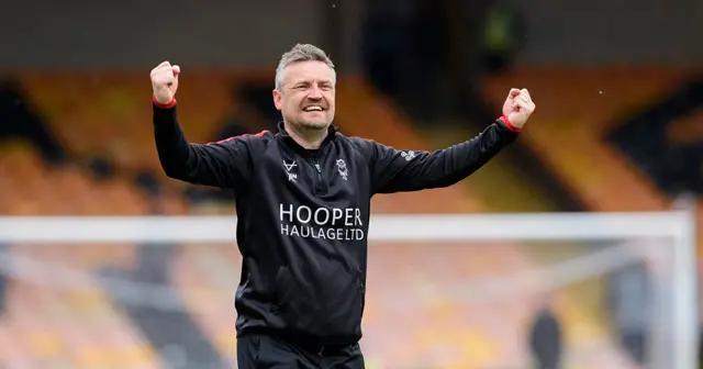 A man wearing a long-sleeve black tracksuit top has both arms aloft with his fists clenched. He is standing on a football pitch with a football goal behind him.