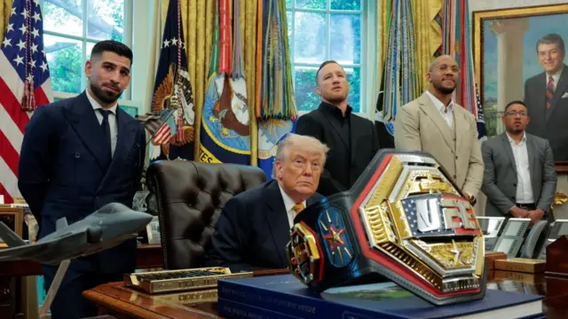Donald Trump sits at the Oval Office desk with the UFC champion belt in front of him and several UFC fighters standing either side of him