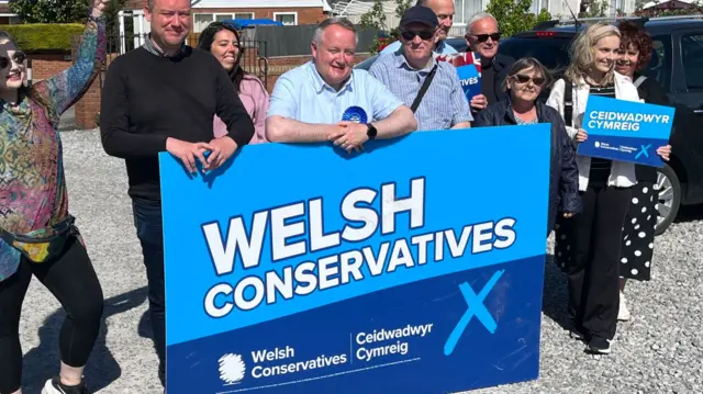 Darren Millar is learning on a large blue sign which says Welsh Conservatives on it. He is surrounded by about a dozen people, some who are holding the same placard but smaller.