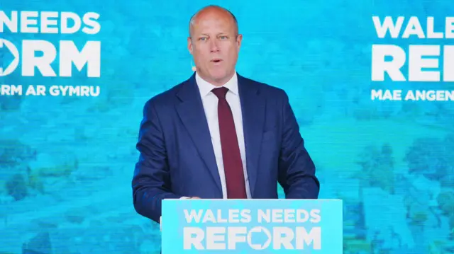 Dan Thomas speaking in front of a screen that says Wales needs Reform, wearing a blue suit and red tie.