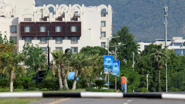 A hotel building seen behind several palm trees at the end of a road. A worker can be seen dusting the street beside signs that say: "Islamabad talks"