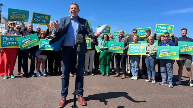 Rhun ap Iorwerth speaks into a microphone standing in front of supporters holding Plaid Cymru placards