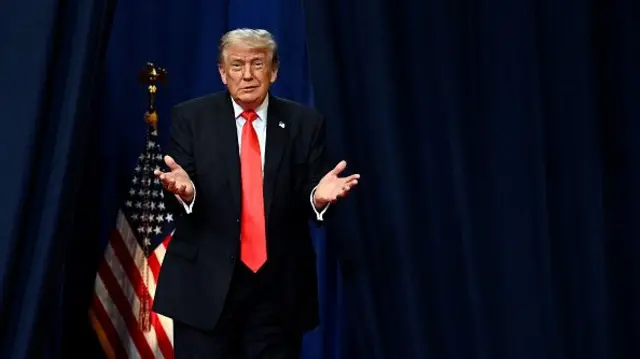 Donald Trump makes a weighing up gesture with his hands, standing on a stage.