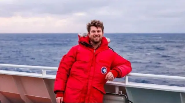 Jake in a large red puffer winter coat leaning against a railing on a boat smiling at the camera in front of the sea