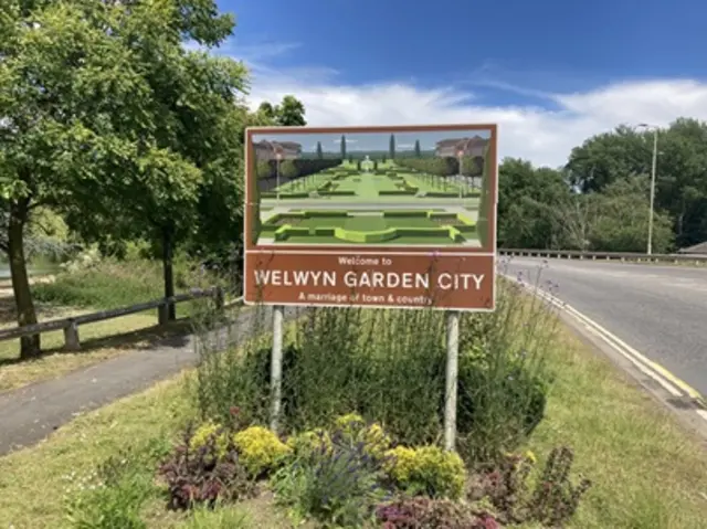 A Welwyn Garden City sign, showing a picture of a Elizabethan knot garden. The sign is standing on a grass verge which hosts some small shrubs. There is a road to the right and a path to the left, lined by trees and a low wooden fence.
