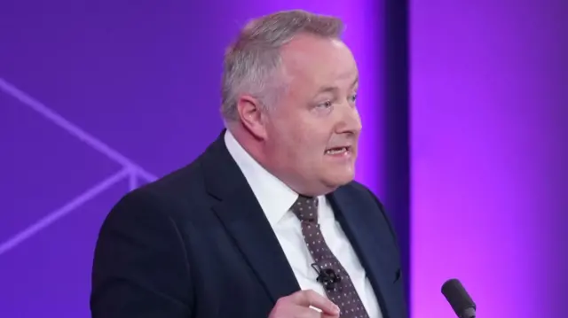 Darren Millar is wearing a suit, tie and shirt. He is standing in front of a purple background, and a podium with a microphone is placed in front of him