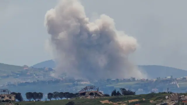 Smoke rises in the distance of a countryside view, with mountains in the background and trees in the foreground