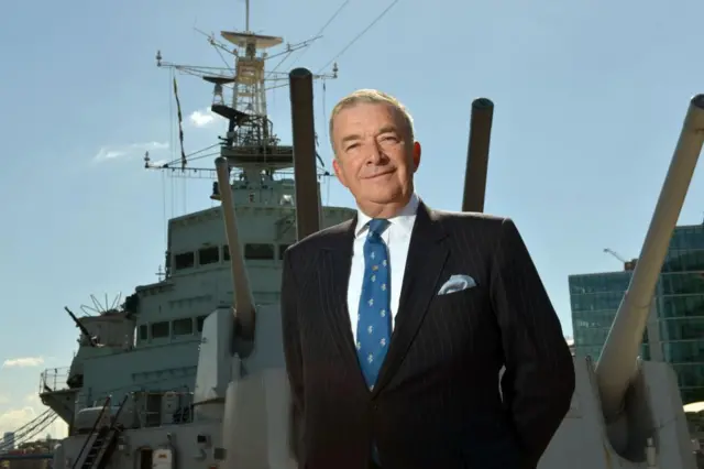 Admiral Lord West is pictured wearing a suit and tie on the deck of a navy ship.