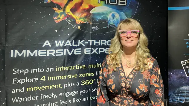 A person stands in front of a colourful exhibition backdrop reading "The Spectrum Place Hub", featuring bright cosmic and abstract imagery. The individual wears pink-framed glasses and a patterned outfit, with text on the display describing a walk-through immersive experience.