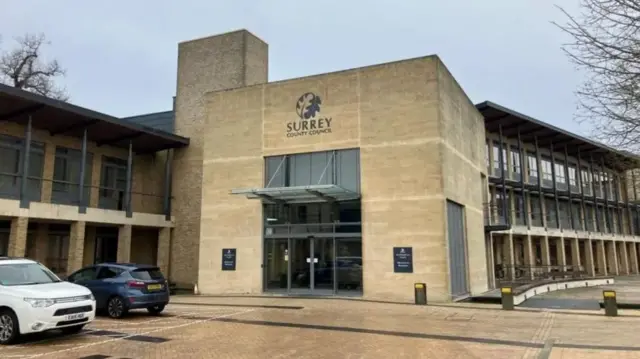 Exterior of Surrey County Council building. A large, light-brown building with the council's logo above the main door at the front. In front and to the left of building in the image are two parked vehicles.