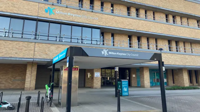 A multi-storey brick office building bearing the sign "MK Milton Keynes City Council", viewed from street level on a clear day. The facade features repeating vertical window recesses and metal railings along each level.