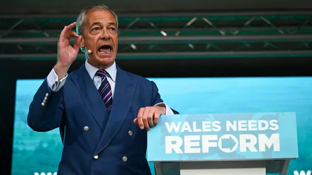 Nigel Farage gestures while standing at a lectern with "Wales needs Reform" written on it
