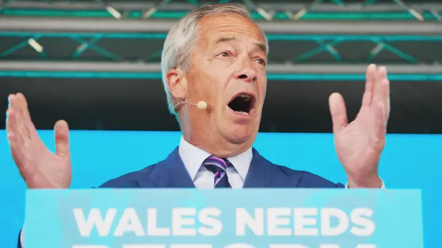 Nigel Farage with his hands outstretched, speaking to a crowd not visible in the photograph.