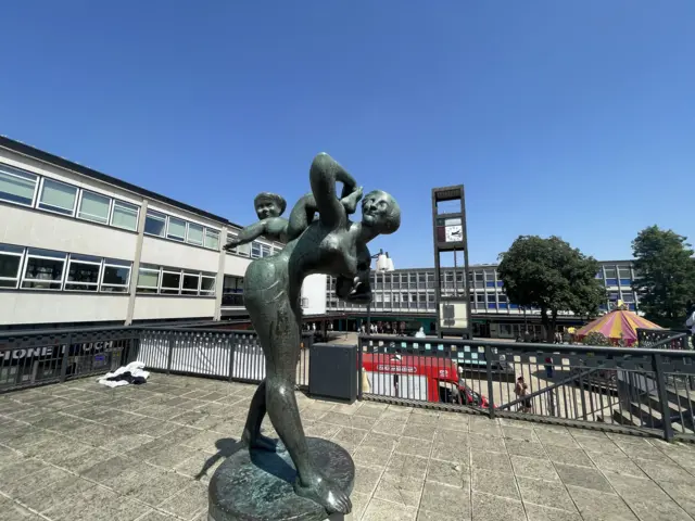 Modern town centre, showing a two-storey concrete building to the left, a tall square segment sign and trees in the background, and a sculpture in bronze of a woman carrying a baby in the foreground.
