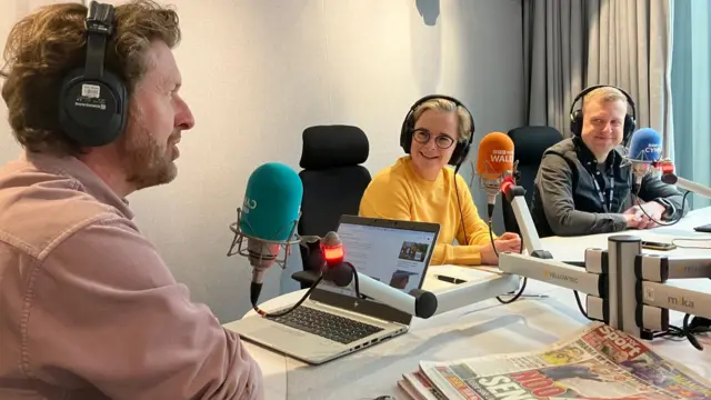 Two men and a woman sat at a studio desk. Felicity Evans is in the middle of James Williams, who is to the left, and Dan Davies, who is to her right. Newspapers can be seen in shot, as well as BBC Wales microphones.