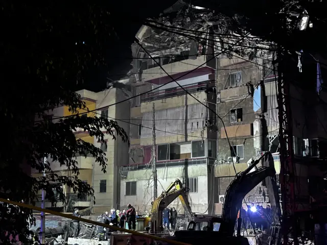 A view from the front of a building destroyed in an Israeli strike. Silhouettes of diggers are in the foreground with emergency workers assessing the damage