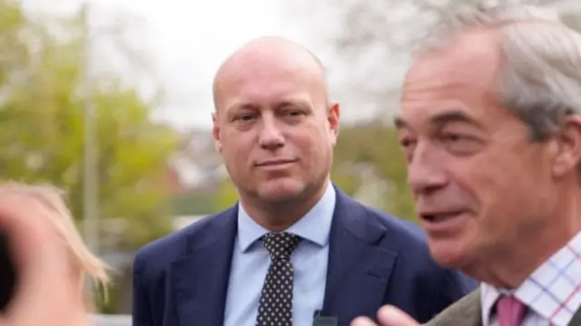Dan Thomas is wearing a suit, tie and shirt while looking at Nigel Farage talking to people.