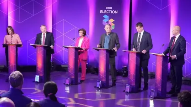 Six leaders stood next to each other in a TV studio in front of lecterns, with the presenter stood to the right.