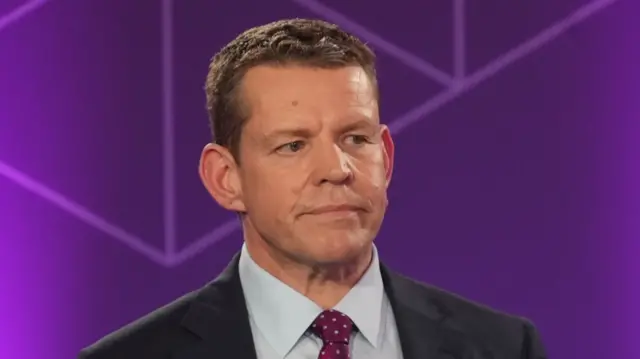 Rhun ap Iorwerth is wearing a suit, tie and shirt and is standing against a purple background
