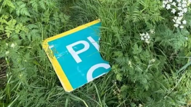 A green and yellow siign, lying in the grass, which has "PL" written on it. A foot away from it is a wooden structure it was attached to.