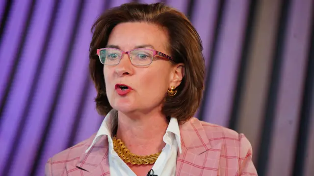 Eluned Morgan is wearing a pink blazer and a gold necklace. She is stood in front of a purple background, wearing glasses.