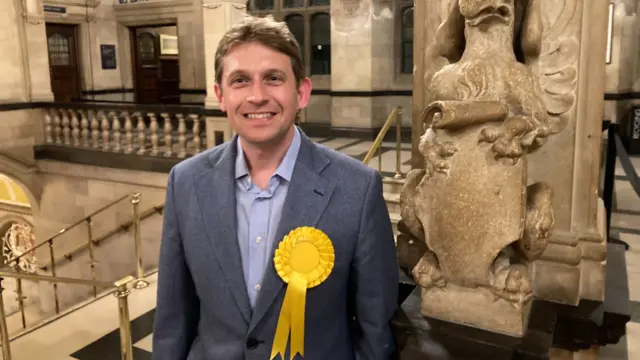 Chris Smowton, Lib Dem leader, wearing a yellow rosette and stood in the town hall in a blue suit smiling at the camera.