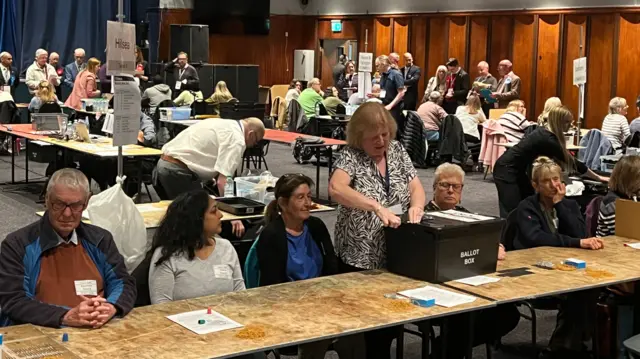 A room full of people sat at wooden tables counting election results.