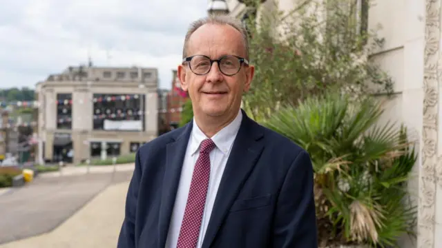 Sir Stephen Houghton stands smiling on a street. He is wearing a suit.