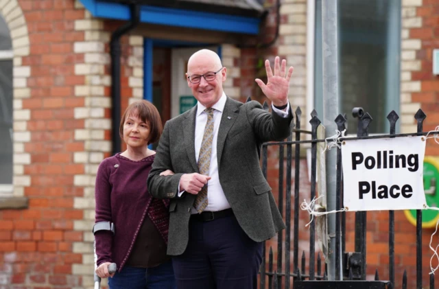 Swinney and Elizabeth wife standing outside a polling station, one raising a hand in greeting beside a "Polling Place" sign
