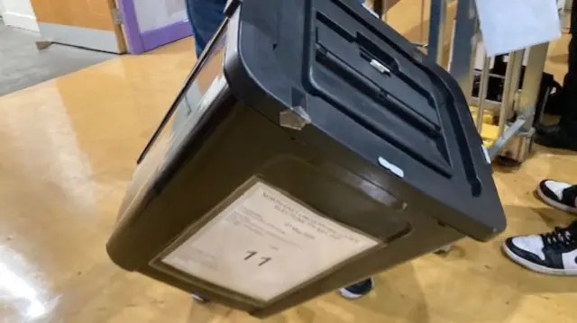 Black box being held in the corner with a white paper sign stuck to the front in a brightly lit room