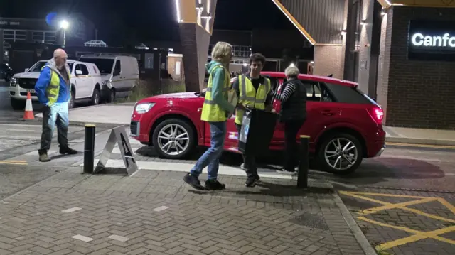 A man in a high-vis jacket is carrying a black box as he steps out of a red car in a sports centre dimly lit with street-lighting