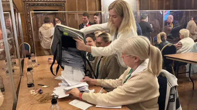 Woman pours a bag of white pieces of paper onto a table with another woman reaching out to catch them