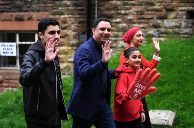 Sarwar with his wife and two boys walking past a polling station sign, raising hands, with one boy holding a large foam hand.