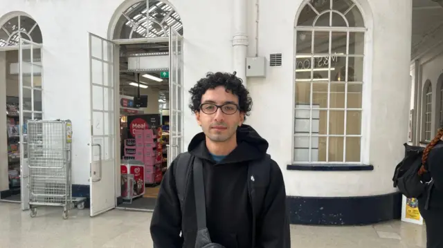 A man in a black hoodie. He is standing outside a white shop in a railway station.