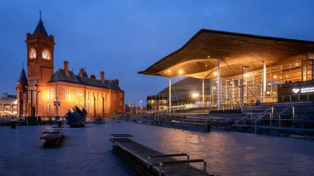 Exterior shot of the Senedd(right), illuminated by lights at night in Cardiff Bay, Cardiff, Wales