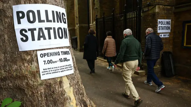 People walking past a tree with a 'Polling Station' sign pinned to a tree
