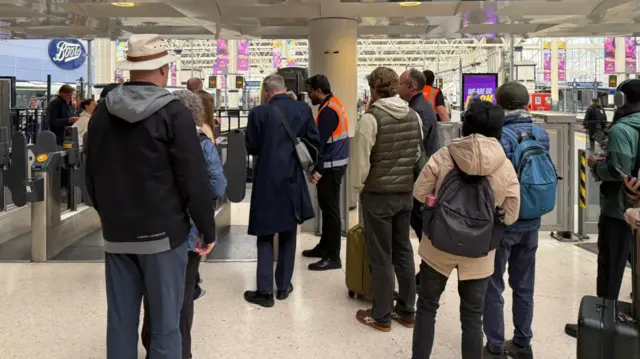 Several people stand near a ticket barrier as staff in hi-vis jackets offer information