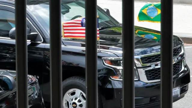 Black car with Brazil and US flags through the black bars of a fence