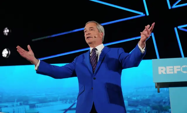 Nigel Farage on a stage. He is wearing a blue suit, a white shirt and a blue and red striped tie. He is wearing a microphone on his cheek and has his arms stretched out to the side. He us standing beside a turquoise lectern with the first four letters of the word Reform on and the outline of a Union flag in blue lights