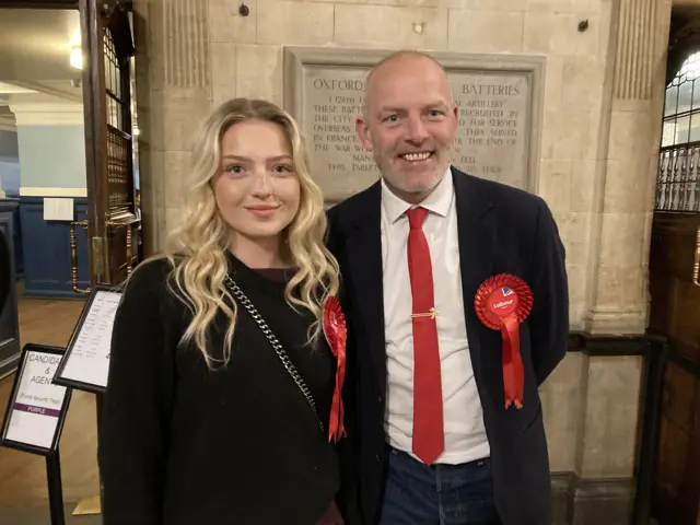 A young woman with blond hair, Emily Lygo, stood next to her father an older man, Mark Lygo. They are both wearing red Labour Party rosettes and stood in a town hall.