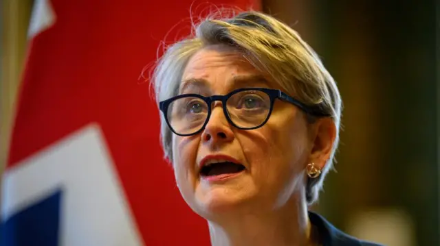 Yvette Cooper stands speaking. A UK flag flies behind her.