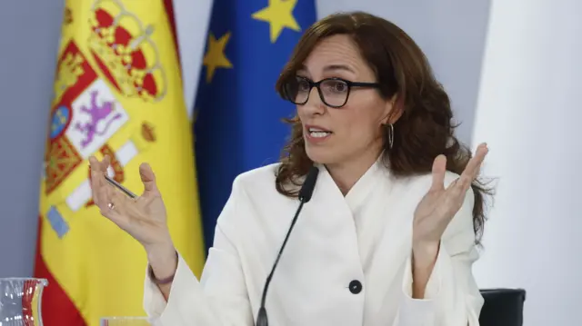 Spanish Minister of Health Mónica García pictured speaking into microphone with white suit and glasses